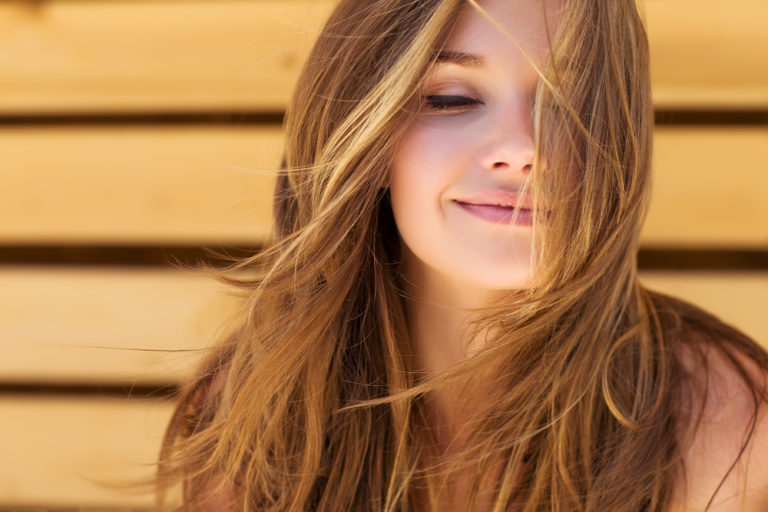 mujer rubia sonriendo con los ojos cerrados pensando con el pelo enla cara y el fondo de madera