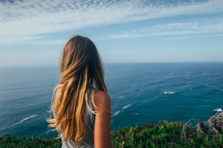 mujer de espaldas dada la vuelta rubia con camiseta de tirantes gris mirando al horizonte al paisaje verde y al cielo azul