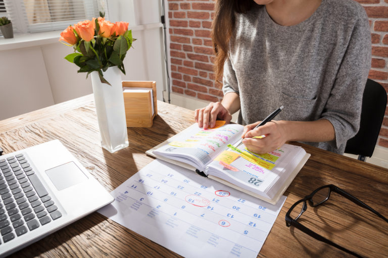 mujer apuntando en una agenda sus tareas y responsabilidades pendientes en una mesa de madera con ordenador al lado y un calendario además de flores y unas gafas