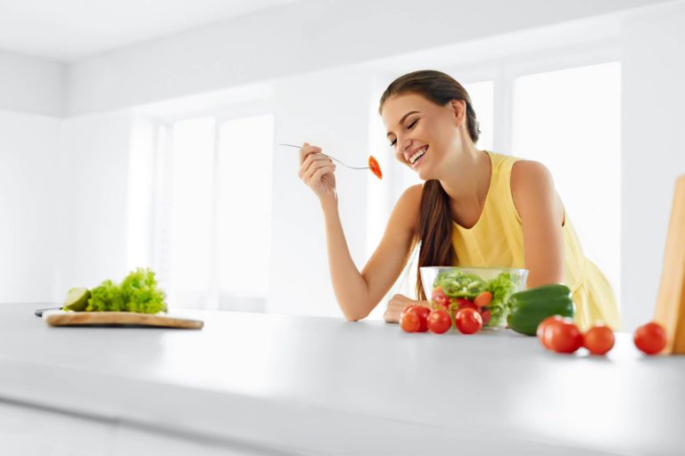 mujer vestida de amarillo comiendo verduras y frutas en cocina blanca y fondo blanco